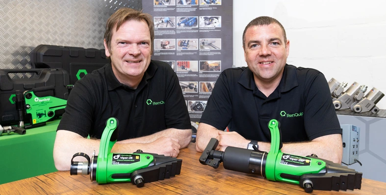 Two men sitting at table with mechanical equipment.